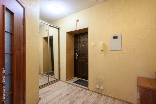 compact apartment hallway features beige textured walls, dark wooden entrance door with panel design, mirrored wardrobe doors. wall-mounted intercom electrical outlets complete functional layout