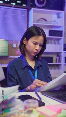 Stressed young Asian office worker feeling overwhelmed with paperwork and workload while working on laptop in office