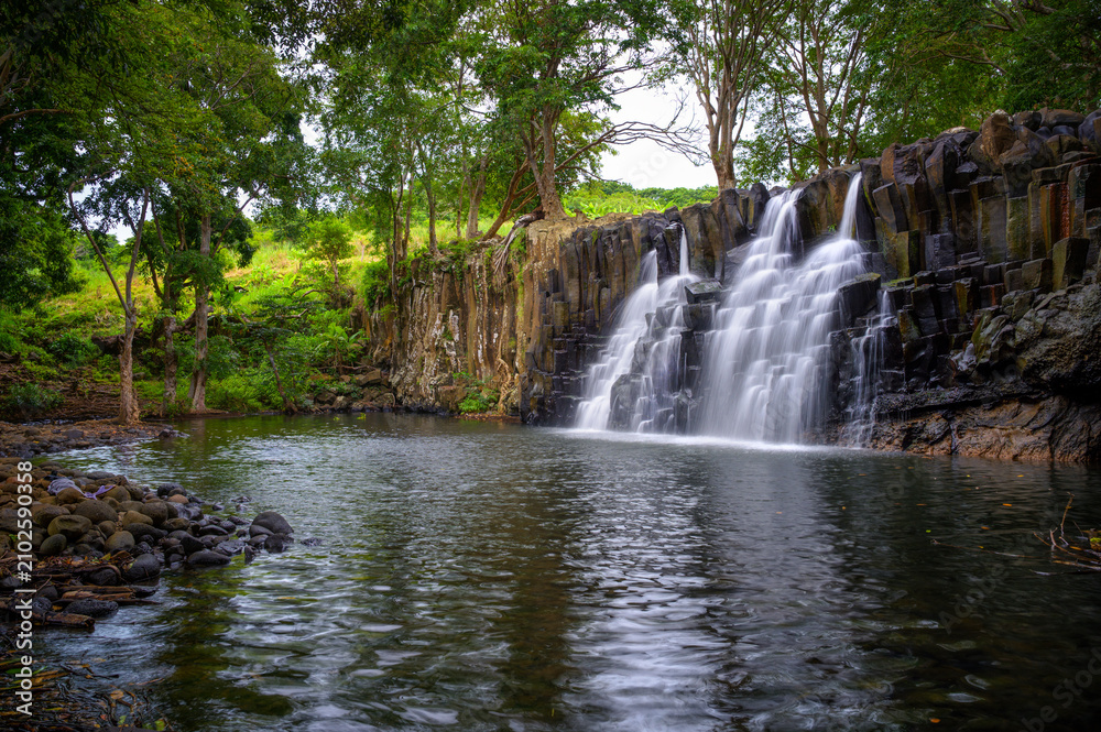 Fototapeta premium Rochester Falls in Mauritius flowing over distinctive basalt rock columns into a calm pool, surrounded by dense tropical forest and lush greenery.