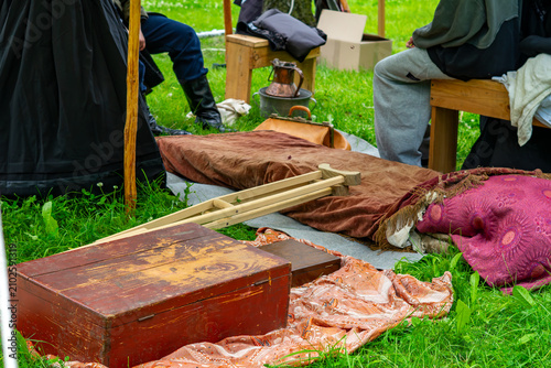 Картината върху платно Close-up of a historical camp with a wooden chest, crutches, brown blanket, decorative pillow, old metal pitcher, and people sitting in period costumes on green grass