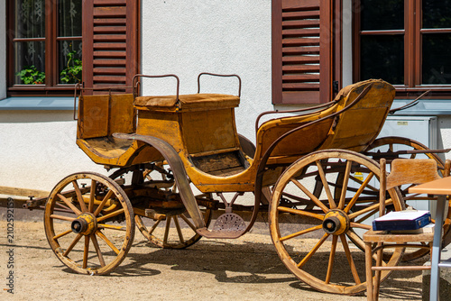 Antique yellow horse-drawn carriage with a wooden frame and large spoked wheels, parked on a sandy surface in front of a building with brown shutters on a sunny day.