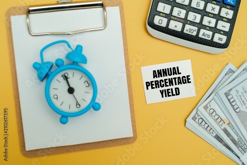 Foto A blue alarm clock on a clipboard with ANNUAL PERCENTAGE YIELD text, calculator and US dollar notes on a yellow background