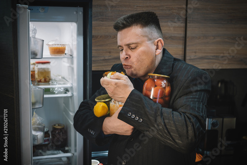 Tableau sur toile Overweight man in a suit eating a sandwich in front of an open fridge, holding jars of food