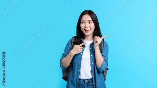 Excited Student with Backpack: A young Asian woman, likely a student, points to the side with a joyful and surprised expression, wearing a backpack and casual attire against a bright blue background.