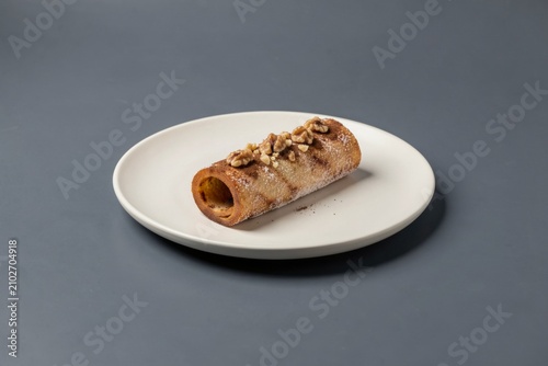 Trdelnik on small white plate against plain grey background. Concept of grilled pastry coated with sugar walnuts, showcasing golden-brown crust and nutty flavor.