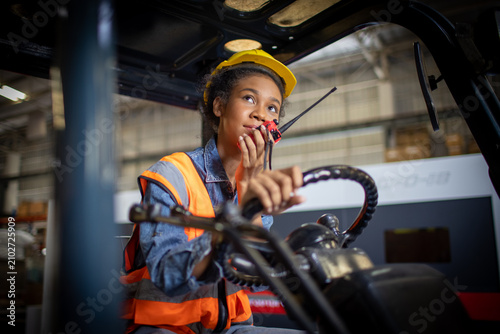 African american female factory worker holding walkie talkie driving forklift in industrial container warehouse storage. Cheerful female logistics worker with hard hat driving forklift truck.