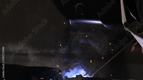 Close up of a welder hands in gloves holding electrode holder with flying sparks in dark workshop