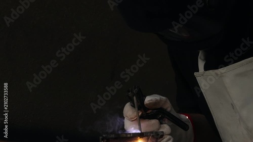 Close up of a welder hands in gloves holding electrode holder with flying sparks in dark workshop