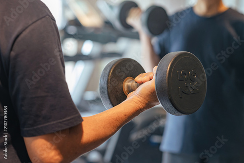 Man gripping dumbbell during workout session in modern gym with training partner nearby, representing social fitness, strength building, muscle recovery and healthy active lifestyle wellbeing