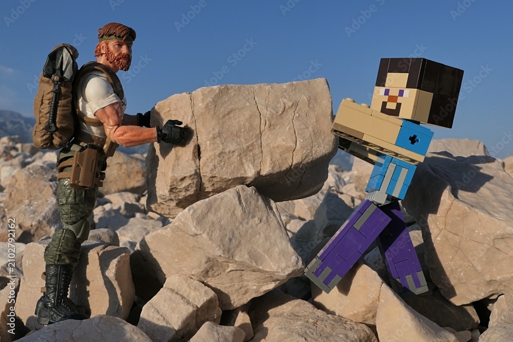 Obraz premium Minecraft action figure of Steve with GI JOE action figure of survival instructor Outback, moving large square shaped coastal rock across the other on Croatian beach, blue summer skies in background. 