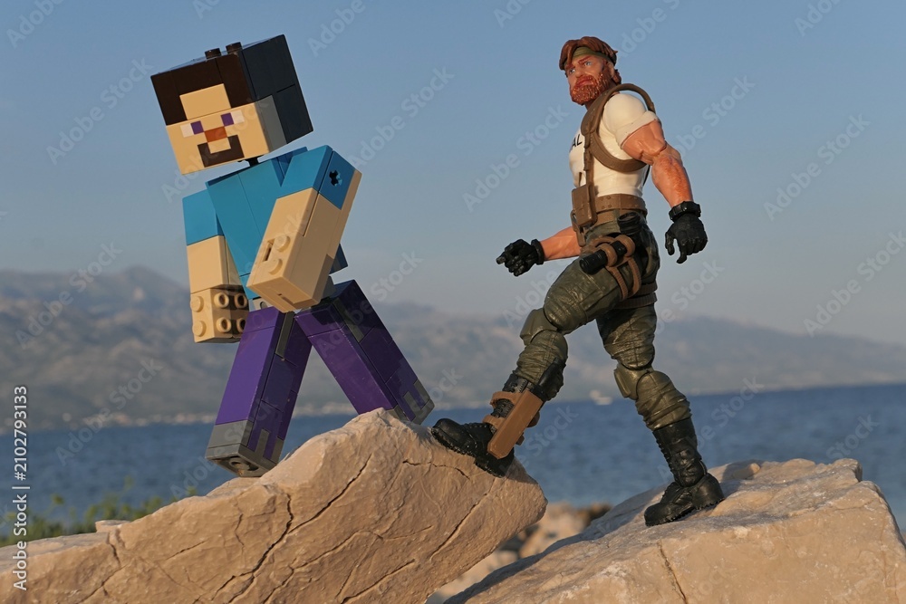 Obraz premium Figures of Minecraft game hero Steve and GI JOE Classified edition survival instructor Outback trekking on sharp coastal rock of Croatian shoreline, bay in Razanac and Velebit mountain in background. 