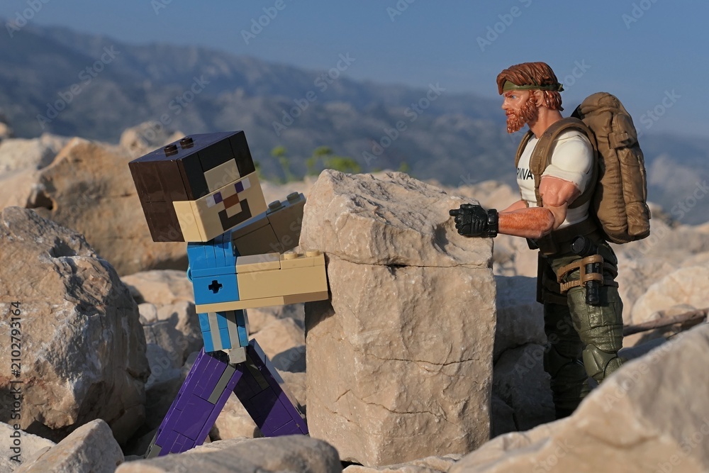 Obraz premium Action figure of Minecraft Steve and GI JOE Classified edtition survival instructor Outback lifting large square block on on Croatian beach, summer daylight sunshine. 