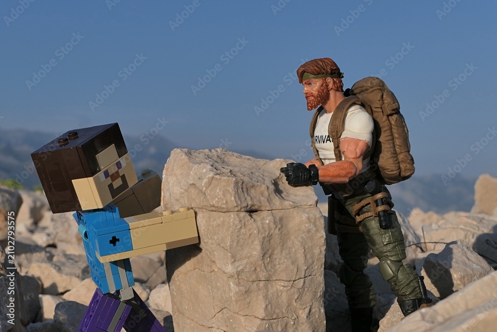 Obraz premium Figures Minecraft Steve and GI JOE Classified edtition survival instructor Outback placing large square block on on Croatian beach, summer daylight sunshine. 