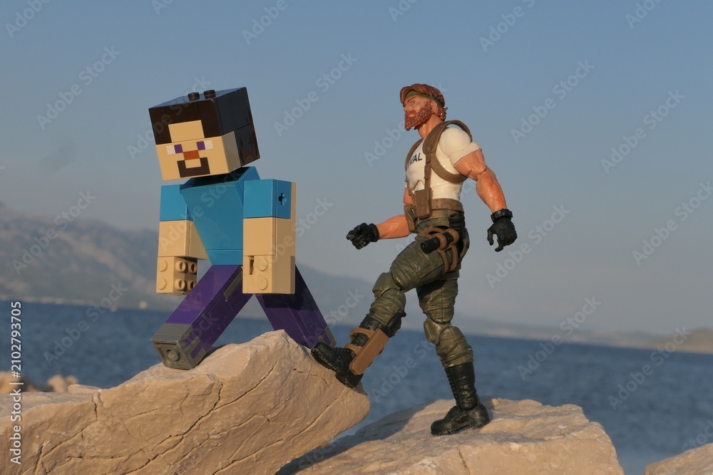 Obraz premium Figures of Minecraft game hero Steve and GI JOE Classified edition survival instructor Outback walking on sharp coastal rock of Croatian shoreline, bay in Razanac and Velebit mountain in background. 