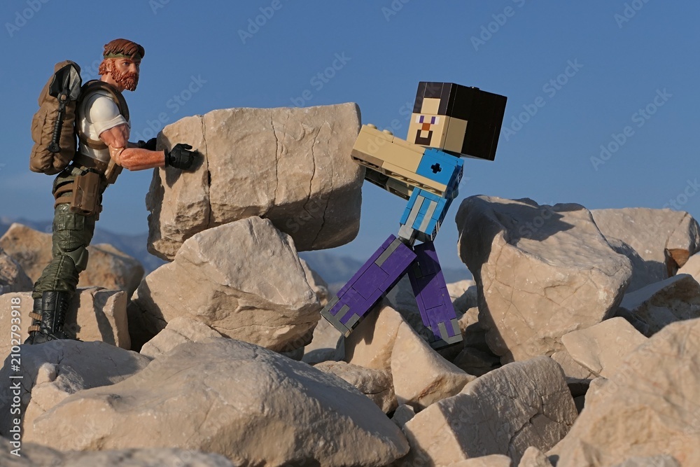 Obraz premium Action figures Minecraft game hero Steve and GI JOE Classified edtition survival instructor Outback placing large square block on on Croatian beach, summer daylight sunshine. 