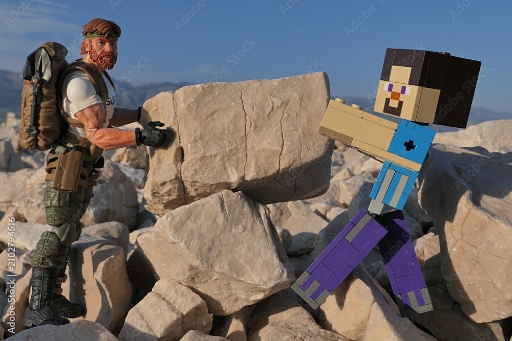 Obraz premium Action figures Minecraft Steve and GI JOE Classified edtition survival instructor Outback placing large square block on on Croatian beach, summer daylight sunshine. 