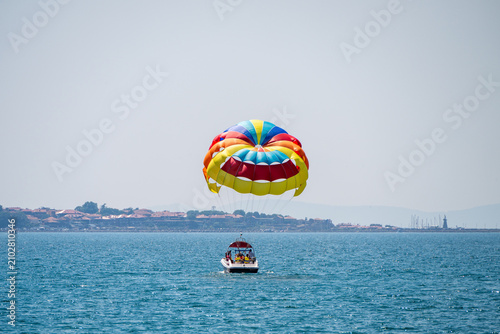 Принти на полотні a vibrant, multi-colored parasail being towed by a motorboat across a sparkling blue sea, with a picturesque coastal town visible along the distant shoreline under a clear sky