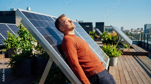 Mature man leaning against solar panel on rooftop, enjoying sunlight and urban cityscape in background, promoting renewable energy.