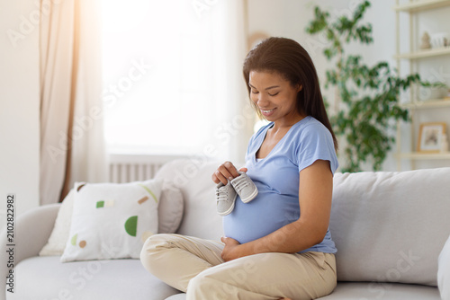 Fotografie Smiling Black pregnant woman holding tiny newborn shoes above her belly while si