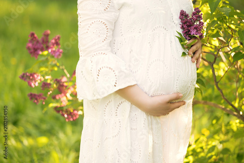 Photography A pregnant woman stands outdoors in a sunny blooming garden, gently cradling her belly
