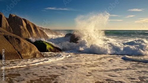 Ocean waves crash against large coastal rocks on a sunny beach