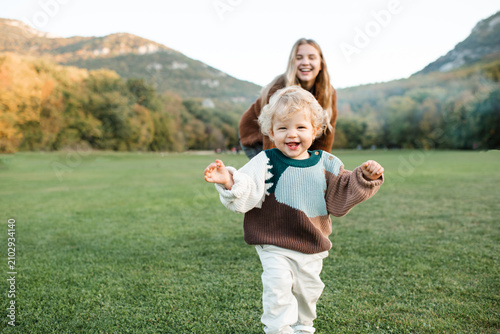 Quadro su tela Smiling little baby girl 1-2 year old running from her mother in green grass lawn and mountain landscape at background