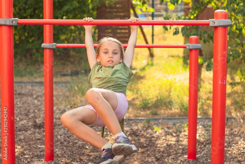 Canvastavla Young girl hanging from red monkey bars in a playground, wearing green shirt and