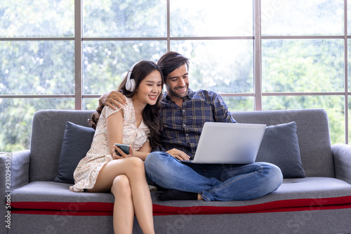 Happy multiethnic couple sitting on sofa and using laptop together at homeCheerful young man and woman watching media on laptop in living roomSmiling Asian woman with headphones and Caucasian man usin