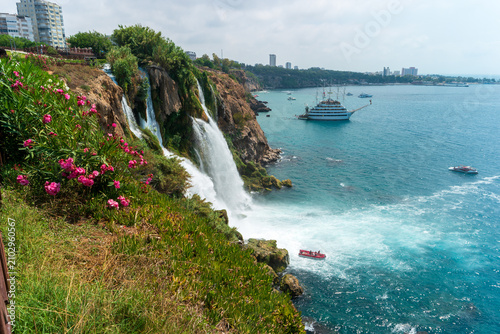 Antalya, Turkey. Lower Duden Waterfall plunging from a flower-covered cliff into the turquoise Mediterranean, with a sightseeing boat below and the modern city skyline beyond