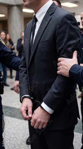 A man in a suit with handcuffed wrists being apprehended by police in a formal indoor setting, conveying a tense atmosphere