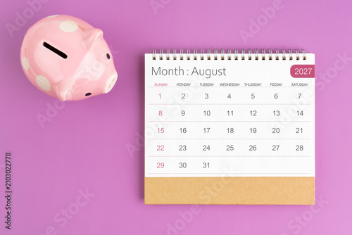 August 2027 desk calendar and pink piggy bank on purple background.