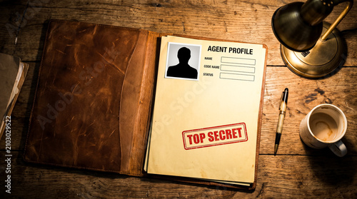 An open file folder labeled Agent Profile with TOP SECRET stamped on it, sits on a wooden desk next to a lamp and a coffee mug