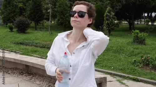woman recovering from cancer holds a water bottle and cools her neck in a city park, illustrating overheating prevention, hydration routine and summer health awareness