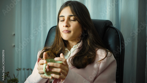 Φωτογραφία Young Hispanic businesswoman taking a break in her office while drinking a cup of coffee, relaxing briefly during her workday in a modern professional workplace