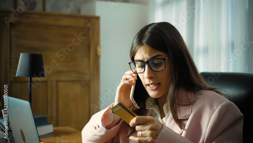 Schilderij op canvas Young Hispanic businesswoman in her office making a phone call about credit card problems, discussing a financial issue and seeking assistance during her workday in a modern workplace