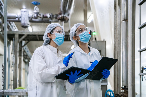 Two workers in white lab coats and face masks stand in an industrial facility. They wear hairnets and blue gloves. One holds a clipboard, the other points with a pen