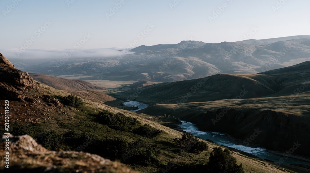 Obraz premium Mist drifts over the vast rugged mountain terrain where a river flows through a quiet green valley