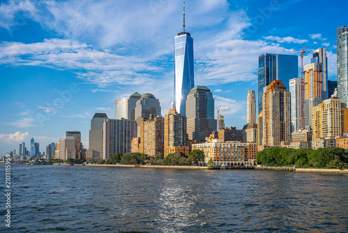 One World Trade Center towering over Manhattan in New York. Sunlight reflects on glass facade of modern skyscrapers along Hudson River.