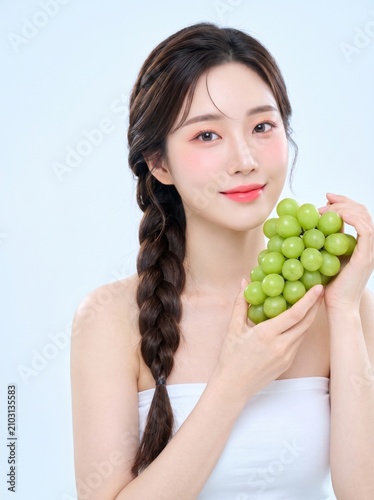 young woman with long dark braided hairstyle, natural makeup, wearing a white strapless tube top, holding fresh green grapes, with a soft smile, isolated on a blue seamless studio background