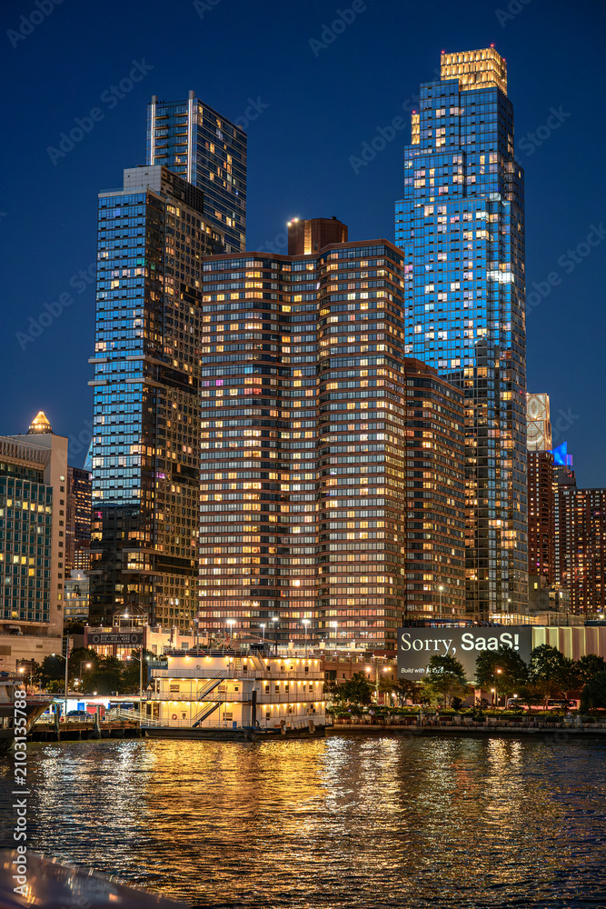 Fototapeta premium New York, USA, 8 June 2026 : Vertical view of New York City skyscrapers illuminated at night. Lit windows of modern apartment buildings reflect yellow light on the river surface, bottom up shot.