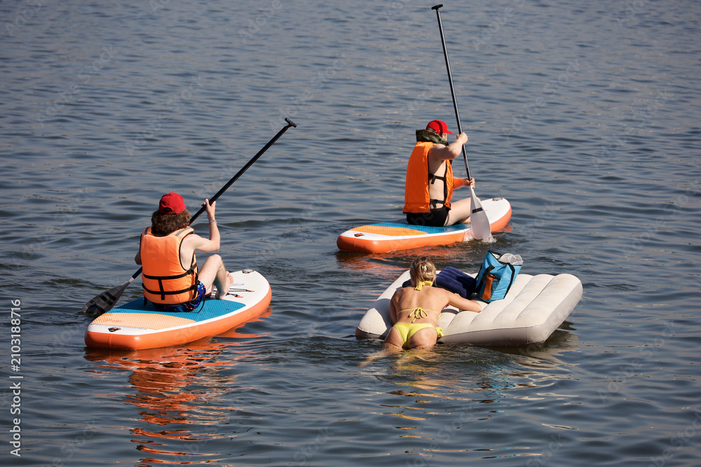 Obraz premium Sup surfing, two guys and girl sailing on a boards and inflatable mattress in water. Stand up paddle boarding in summer