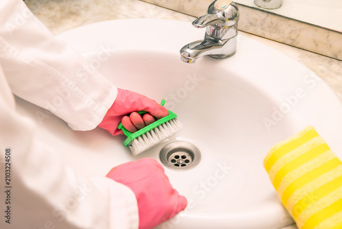 Person wears a white lab coat and pink gloves. They clean a white ceramic sink with a green brush. The brush has pink scrubbers and white bristles. Chrome faucet and drain sit in the sink