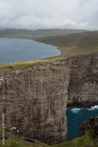 Massive cliffs of Trælanípa drop into the Atlantic Ocean beside Lake Sørvágsvatn, with a small boat crossing the lake and the village of Miðvágur visible in the distance, Vágar, Faroe Islands