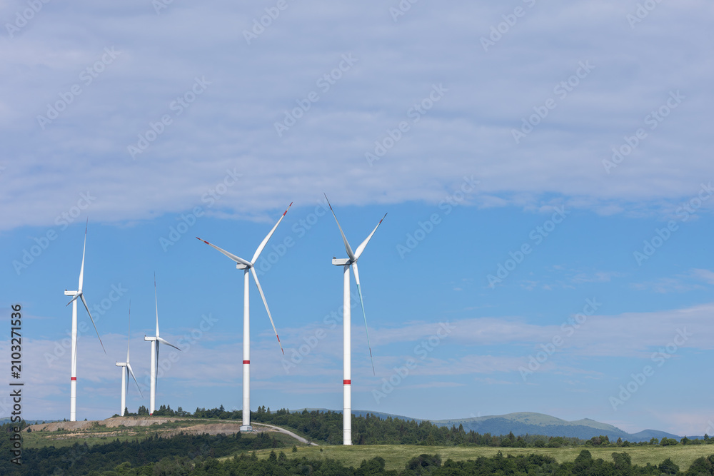 Fototapeta premium A view of multiple large white wind turbines on a green hill, with rolling mountains and a blue sky in the background with Copy space