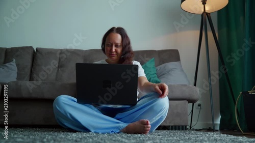 Woman sitting cross legged on carpet floor using laptop computer in contemporary living room with gray sofa, tripod lamp, green curtains, calm indoor setting.