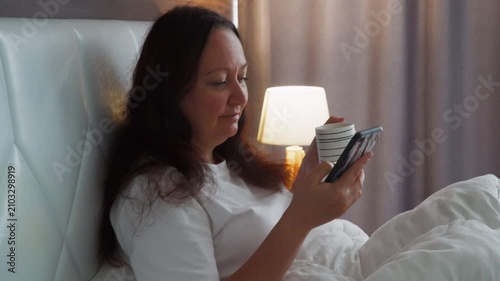Woman in white shirt sits on bed holding smartphone, backlit by warm table lamp, peaceful evening moment in comfortable bedroom setting.