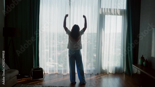 Silhouetted woman standing at floor to ceiling window with arms raised in celebration, overlooking expansive city skyline from luxury high rise apartment, backlit natural light through sheer curtains.