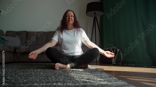 Woman in white shirt seated cross legged on carpet stretches both arms overhead in relaxed pose inside modern apartment with green curtain, sofa, wooden floor
