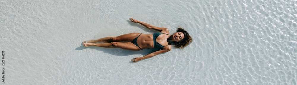 Fototapeta premium Top view of woman lying on rippled white sand in black bikini for summer background and wellness banner