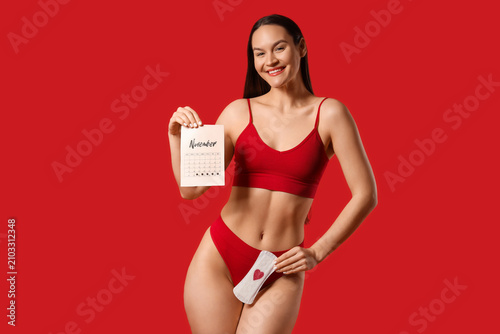 Young woman in underwear with menstrual calendar and pad on red background